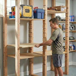 May include: A man installing a wooden storage shelf in a garage. The unfinished wooden structure has multiple shelves, with various tools and containers on the shelves. A yellow toolbox and blue container are visible.