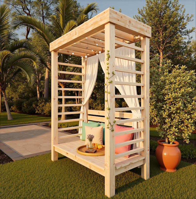 May include: A wooden outdoor gazebo with a slatted roof and sides, draped with white curtains. The gazebo has a built-in bench with colorful pillows and a tray with a vase and fruit. String lights are strung across the top. The gazebo is set on a green lawn.