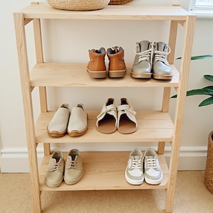 May include: A wooden shoe rack displaying various pairs of shoes. The shoes include brown leather boots, gray lace-up boots, beige slip-ons, cream sandals, and beige sneakers with white soles. The rack has four shelves and is made of light-colored wood.
