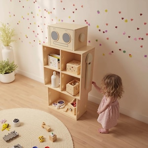 Puede incluir: Una unidad de almacenamiento de madera de color claro con múltiples estantes cúbicos, coronada con una cabeza en forma de caja con recortes circulares. Un niño pequeño con un vestido rosa se acerca a la unidad. La habitación tiene una paleta de colores neutros con confeti en forma de corazón.