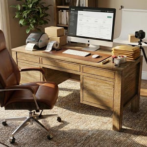 Puede incluir: Una oficina en casa con un escritorio de madera, una silla de oficina de cuero marrón y un ordenador que muestra un sitio web. Hay una impresora de etiquetas, una cámara y equipos de iluminación, lo que sugiere un negocio de comercio electrónico.