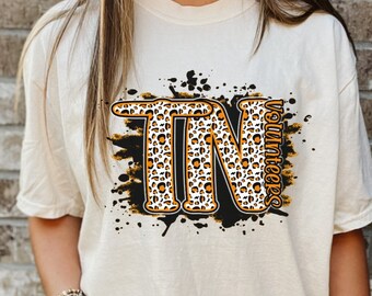 May include: A white t-shirt featuring a black and orange graphic of the letters "TN" in a leopard print design. The letters are surrounded by black paint splatters and the word "Volunteers" is written vertically to the right of the letters.