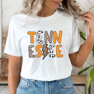 Può includere: Maglietta bianca con la parola "TENNESSEE" in lettere arancioni con un motivo leopardato e una grafica di fulmine.