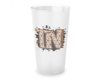 May include: A white frosted glass with a black and orange graphic of the letters "TN" with a leopard print pattern. The letters are surrounded by black paint splatters. The text "Volunteers" is written vertically on the right side of the graphic.