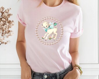 Peut inclure: Un t-shirt rose avec un agneau blanc portant un nœud bleu et un papillon sur le dos. L'agneau est entouré d'un cercle de perles.