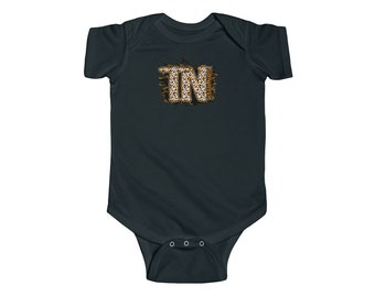 Peut inclure: Une grenouillère bébé gris foncé avec un motif "TN" doré et blanc sur le devant. Le motif a un aspect vieilli et est entouré d'un motif de projection blanche.