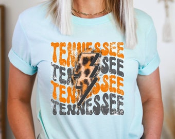 Puede incluir: Camiseta azul claro con mangas enrolladas. Presenta la palabra "Tennessee" en letras bloque naranja y gris, con un rayo con estampado de leopardo en el centro. Una camiseta casual para el día a día.
