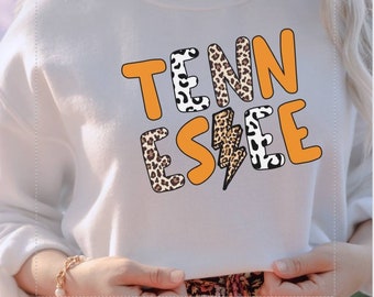 May include: White sweatshirt featuring the word "TENNESSEE" in orange, leopard print, and monochrome lettering. A lightning bolt graphic is integrated into the design. The sweatshirt is held by a person.