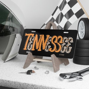 May include: A black license plate with the word "Tennessee" in orange letters with white polka dots and a leopard print border. The license plate is on a wooden stand.