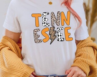 May include: A white t-shirt featuring an orange and black graphic that reads "TENNESSEE" with a lightning bolt in the centre. The graphic has a leopard print pattern.