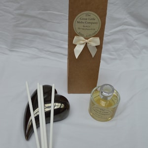 May include: A brown cardboard box with a white ribbon and a label that reads "The Great Little Melts Company Black Pomegranate Reed Diffuser". A clear glass bottle with a silver lid and a label that reads "The Great Little Melts Company Black Pomegranate Reed Diffuser" is next to the box. A black ceramic heart-shaped diffuser base with white reeds is in the background.
