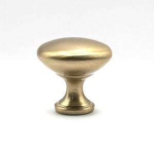 May include: A mushroom-shaped cabinet knob with a brushed gold finish. The hardware features a smooth, rounded top surface that tapers down into a narrow, cylindrical neck and a circular base, creating a solid, metallic furniture component.
