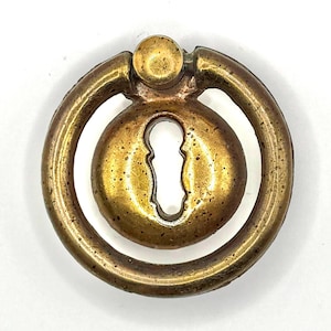 Könnte beinhalten: Ein Messing-Schlüsselloch-Schubladengriff mit einem runden Ringgriff. Der Griff hat ein Schlüsselloch in der Mitte und einen erhabenen Knopf oben.