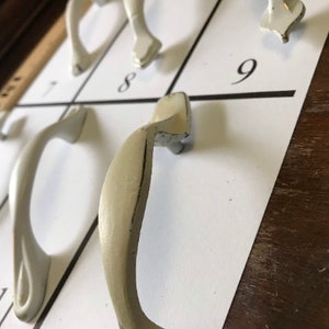 Puede incluir: Una colección de tiradores de armario blanquecinos dispuestos sobre una superficie blanca con números negros. Los tiradores varían en diseño, con formas curvas y angulares. El fondo incluye una superficie de madera y una regla.