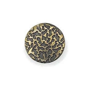 May include: A circular button featuring a metallic gold finish with a recessed, dark gray, irregular organic pattern across its surface. The design consists of interconnected, web-like shapes that create a textured, high-contrast appearance.