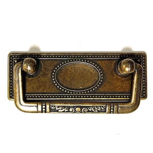 Peut inclure: Poignée de tiroir en laiton antique avec une plaque rectangulaire et un ovale décoratif au centre. La poignée a une poignée en boucle avec des extrémités arrondies.