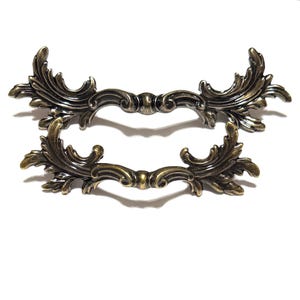 May include: Two antique bronze-colored drawer pulls with ornate, symmetrical designs. Each pull features a curved handle with decorative flourishes and a central connecting element. The pulls are set against a white background.
