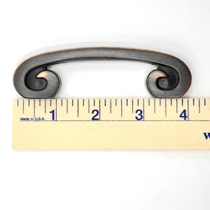 Pode incluir: Puxador de arm&aacute;rio bronzeado com uma al&ccedil;a curva e extremidades enroladas. O puxador mede aproximadamente 10 cm de comprimento, conforme indicado pela r&eacute;gua. Este hardware decorativo &eacute; adequado para gavetas ou arm&aacute;rios.