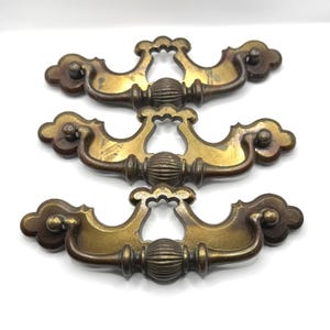 May include: Three antique brass drawer pulls with a decorative design. Each pull features a central, ribbed handle and ornate, cloud-shaped ends. The pulls have a warm, aged patina, suggesting vintage or retro style.