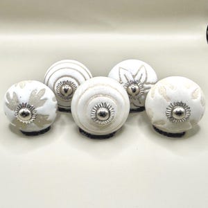 May include: Five round ceramic cabinet knobs with a white base and light beige patterns. Each knob features a central silver-colored metal flower-shaped accent. The designs include floral motifs, concentric circles, and leaf-like shapes on the surface.