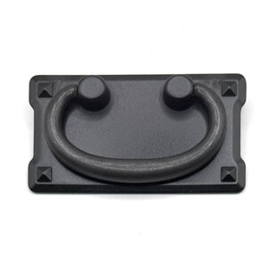 Puede incluir: Un tirador de cajón rectangular en color negro mate que presenta un asa central oscilante unida a una placa posterior plana. La placa tiene esquinas redondeadas y cuatro pequeños pernos en forma de pirámide en cada esquina.