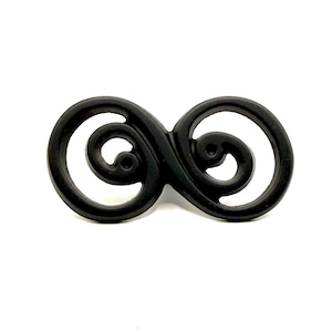 Puede incluir: Un broche de metal negro mate con forma de símbolo de infinito y dos espirales circulares. El diseño presenta un marco liso y continuo que se curva en dos espirales distintas, creando un patrón calado con un acabado oscuro y uniforme.