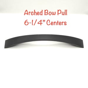 May include: A matte black arched bow pull handle with a curved design. The text above the handle reads Arched Bow Pull 6-1/4" Centers, indicating the distance between the mounting points in inches for this cabinet hardware piece.