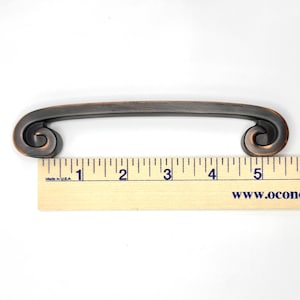Pode incluir: Um puxador de arm&aacute;rio bronzeado com uma pega curva e extremidades decorativas em espiral. O puxador mede aproximadamente 12.7 cm de comprimento, conforme indicado pela r&eacute;gua. Ideal para m&oacute;veis e arm&aacute;rios.