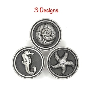 May include: Three circular metal buttons with a dark gray background and silver-toned raised relief designs. The designs include a spiral seashell, a seahorse, and a starfish. The text 3 Designs is printed in red above the buttons.