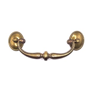 May include: A brass-toned metal drawer pull featuring a curved, swinging handle attached to two oval-shaped backplates. Each backplate is decorated with a rope-style border, and the central handle has a smooth, contoured finish with a central node.