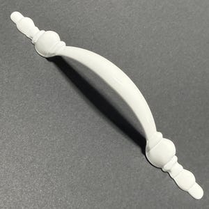 Op de afbeelding: Een witte kastgreep met een gebogen handvat en decoratieve uiteinden. De hardware is gemaakt van een glanzend materiaal en is ontworpen voor een gemakkelijke grip. De greep wordt getoond tegen een grijze achtergrond.
