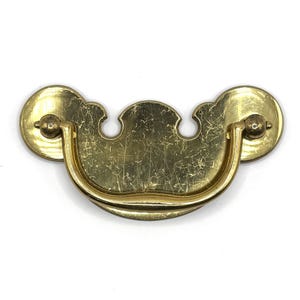May include: A brass drawer pull featuring a wide, curved backplate with a scalloped top edge and a central, U-shaped swinging handle. The metal surface displays visible surface scratches and a polished gold-toned finish throughout the hardware.
