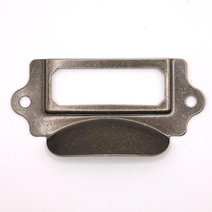 Puede incluir: Portaetiquetas con acabado en bronce antiguo, con una ventana rectangular para la etiqueta y un asa curvada. El soporte tiene dos pequeños agujeros a cada lado para el montaje. El metal tiene un acabado envejecido, lo que le da un aspecto vintage.
