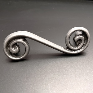 Puede incluir: Un tirador de armario de metal plateado con un diseño decorativo de espiral. El tirador tiene un acabado liso y ligeramente texturizado.