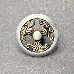 May include: Circular silver and gold-toned button with an ornate, swirling design. The button has a central, raised, circular element and a decorative pattern within a circular frame. Likely for clothing or crafts.