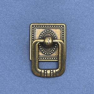 May include: A gold-toned metal cabinet pull with a square base and a ring handle. The base has a decorative pattern of small dots and circles.