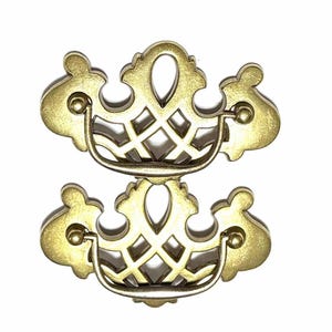 Peut inclure: Deux poignées de tiroir en laiton antique avec un design ajouré orné. Chaque poignée présente une poignée incurvée et des ornements décoratifs, avec une forme ovale centrale et un treillis en losanges. Le matériel est sur fond blanc.