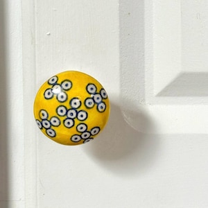 May include: A yellow ceramic knob with a blue and white pattern of circles. The knob is round and has a smooth surface.