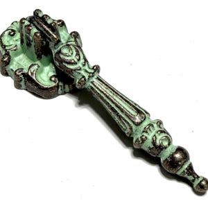 May include: A decorative cabinet pull in a distressed mint green and bronze finish. The handle has an antique style with intricate detailing and a long stem. Suitable for drawers, cabinets, or furniture restoration. The pull measures approximately 15 cm in length.