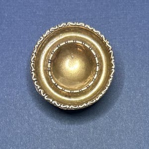 May include: A gold-coloured, circular object with a decorative edge. The object has a bowl-like shape with concentric rings and a detailed, ornate design. The centre is a slightly raised, rounded shape.