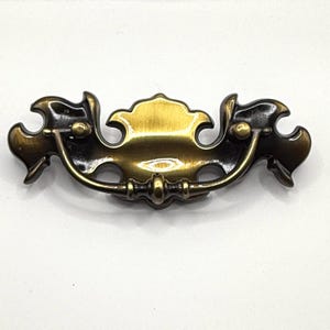 May include: A decorative brass cabinet pull with a scrolled design. The pull has a dark brown finish and a gold center.