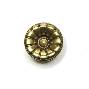 May include: A round, antique-style button with a gold finish. The button has a central raised circle surrounded by a series of petal-like shapes. The button is likely made of metal and has a slightly tarnished appearance.