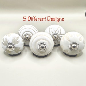 May include: Five round ceramic cabinet knobs with white surfaces and beige patterns. Each knob features a central silver-toned metal hardware piece. The text 5 Different Designs is printed in red above the knobs, which are arranged in a group.