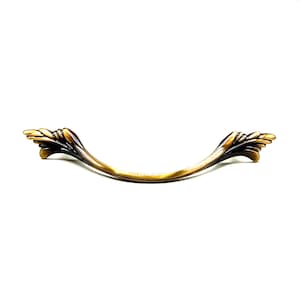 May include: A curved drawer pull handle featuring a metallic antique brass finish with dark brown shading in the recessed areas. The handle has decorative, wing-like ends with layered, leaf-inspired detailing on both sides of the central bar.