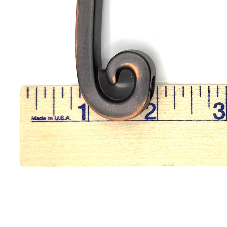 Pode incluir: Close-up de um puxador de arm&aacute;rio de cor bronze com um design decorativo de rolagem. O puxador tem aproximadamente 5 cm de comprimento e &eacute; fabricado nos EUA. O puxador est&aacute; em um fundo branco.