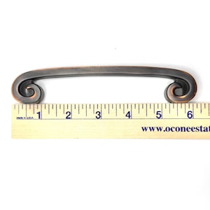 Pode incluir: Puxador de arm&aacute;rio em tom bronze com uma al&ccedil;a curva e design de rolagem em cada extremidade. O puxador mede aproximadamente 15 cm de comprimento. O hardware &eacute; mostrado em uma r&eacute;gua de madeira para escala.