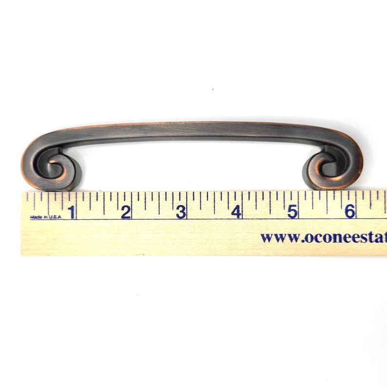 Pode incluir: Puxador de arm&aacute;rio em tom bronze com uma al&ccedil;a curva e design de rolagem em cada extremidade. O puxador mede aproximadamente 15 cm de comprimento. O hardware &eacute; mostrado em uma r&eacute;gua de madeira para escala.