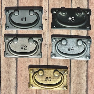 May include: Five different styles of cabinet pulls, each with a different finish. The pulls are rectangular with a rounded arch shape. The finishes include black, silver, and gold.
