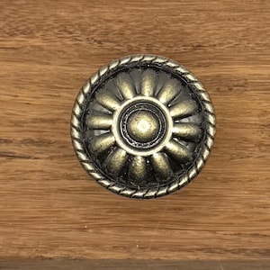 May include: A bronze-colored cabinet knob with a floral design. The knob has a central circular element surrounded by petal-shaped details and a rope-like border. The knob is set against a wooden surface.