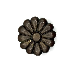 May include: A bronze-coloured, flower-shaped decorative object. The object has a central circular element surrounded by teardrop-shaped petals. The metal has a slightly aged appearance, with hints of gold and green. The object is set against a white background.